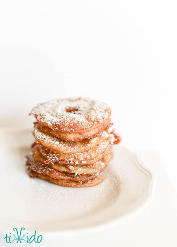 Apple fritter rings dusted with powdered sugar and stacked on a white plate.