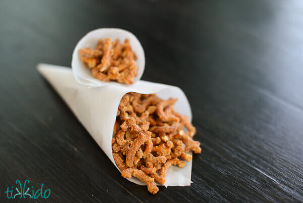 Two parchment paper cones filled with Malted Pretzel Snack Mix on a dark wood surface.