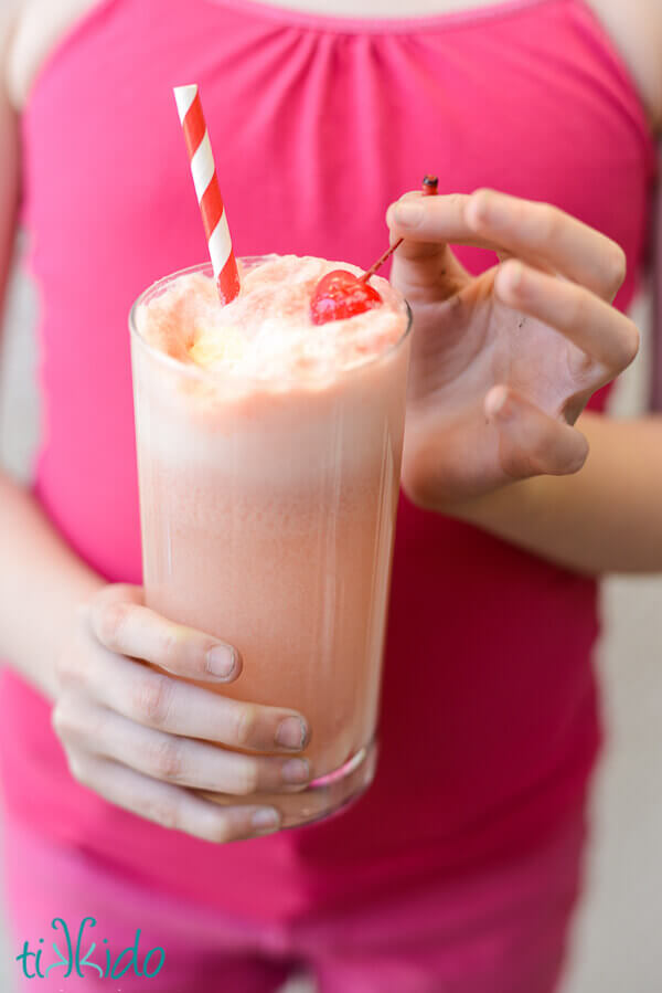 Little girl's hands holding a Shirley Temple Ice Cream Float with a cherry and red and white straw.
