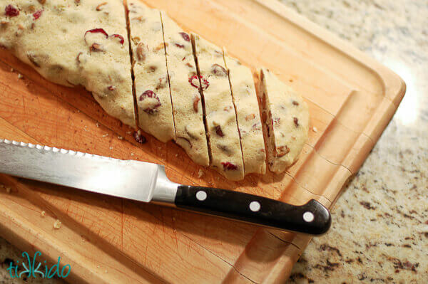 Cranberry Pistachio Biscotti Recipe being cut into slices with a serrated knife on a cutting board.