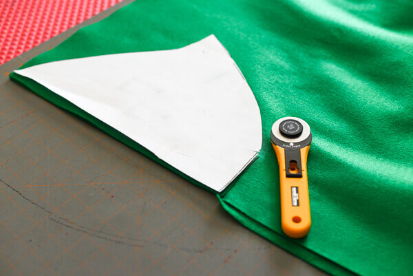 Template of peter pan hat on top of green felt on a self-healing cutting mat, with a yellow rotary fabric cutter.