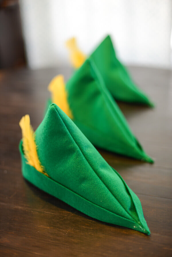 Set of three green felt peter pan hats with yellow feathers, lined up on a dark brown wood table