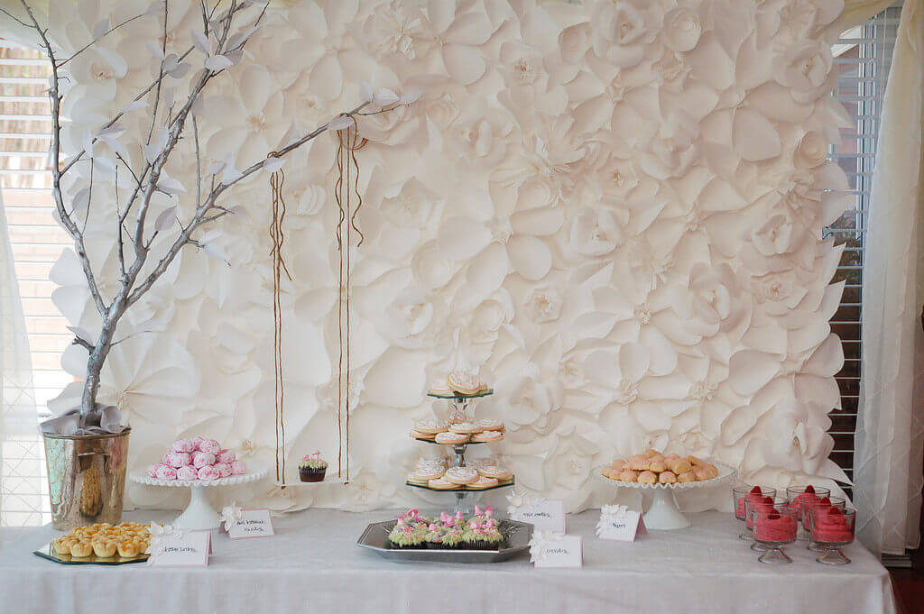 Monochromatic white paper flower backdrop to a dessert table featuring a three tiered serving tray, a silver tree, and a cupcake on a swing.