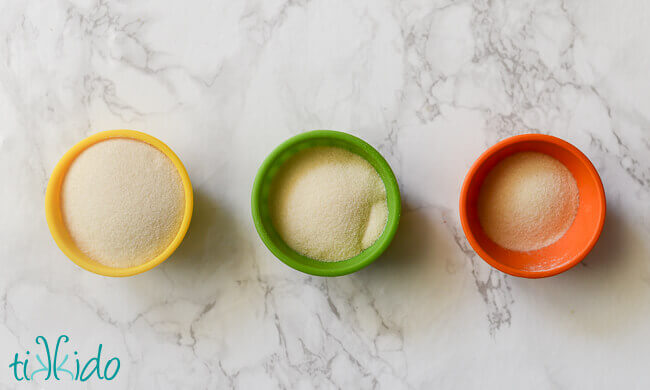 Three small bowls, one yellow, one green, one orange, with varying levels of unflavored Knox gelatin on a white marble background.