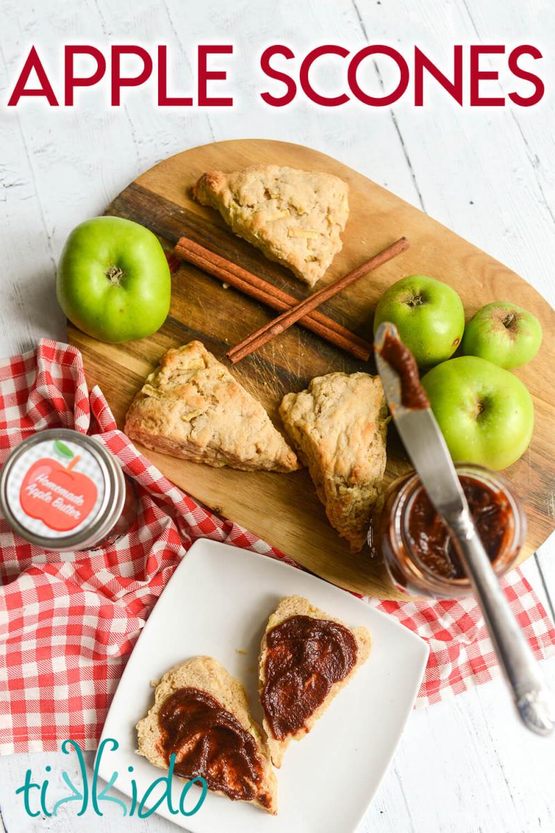 Apple scones on a cutting board, surrounded by fresh apples, cinnamon sticks, and jars of homemade apple butter.  A white plate with an apple scone sliced in half and spread with apple butter sits on a red gingham cloth next to the cutting board.  Text overlay reads "apple scones."