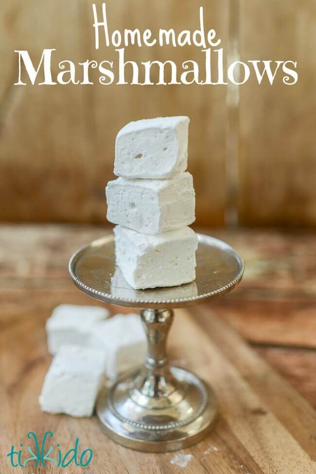 Three homemade marshmallows stacked on a tiny serving tray, and three more homemade marshmallows on the wooden surface below.