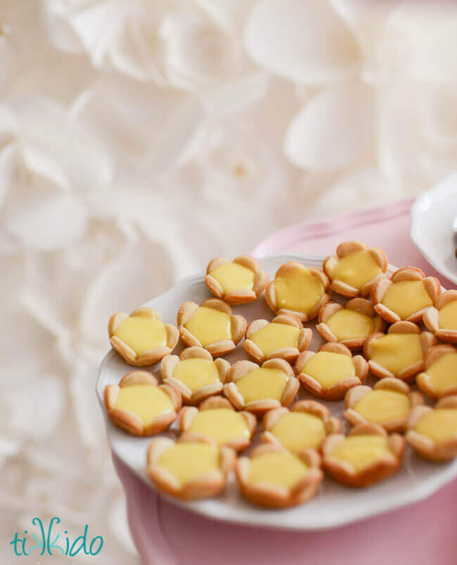 Mini lemon tarts filled with lemon curd on a white plate, in front of a textured paper flower backdrop.