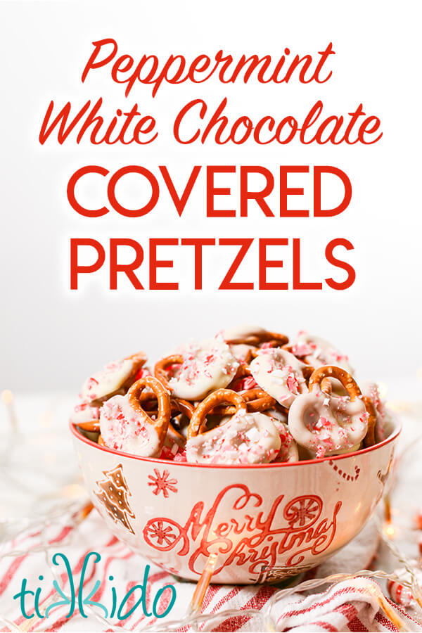 Bowl full of pretzels dipped in peppermint white chocolate and sprinkled with crushed peppermint candies.  Whole pepperming candies, twinkle lights, and a red and white striped kitchen towel surround the bowl.  Text overlay reads "Peppermint White Chocolate Covered Pretzels."