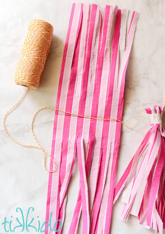 Pink paper napkin cut into fringe for tassel garlands, next to a spool of yellow and white baker's twine.