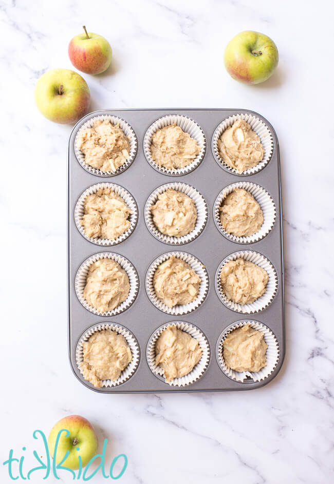 Apple muffin batter, unbaked, in a lined muffin tin, surrounded by fresh apples.