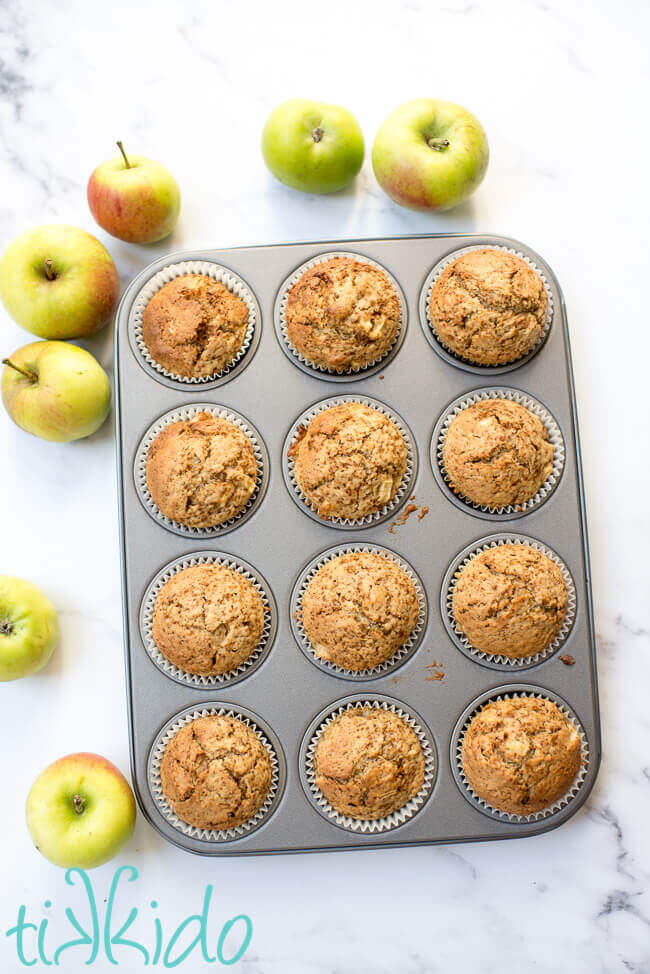 Freshly baked apple muffins in a muffin tin, surrounded by fresh apples.