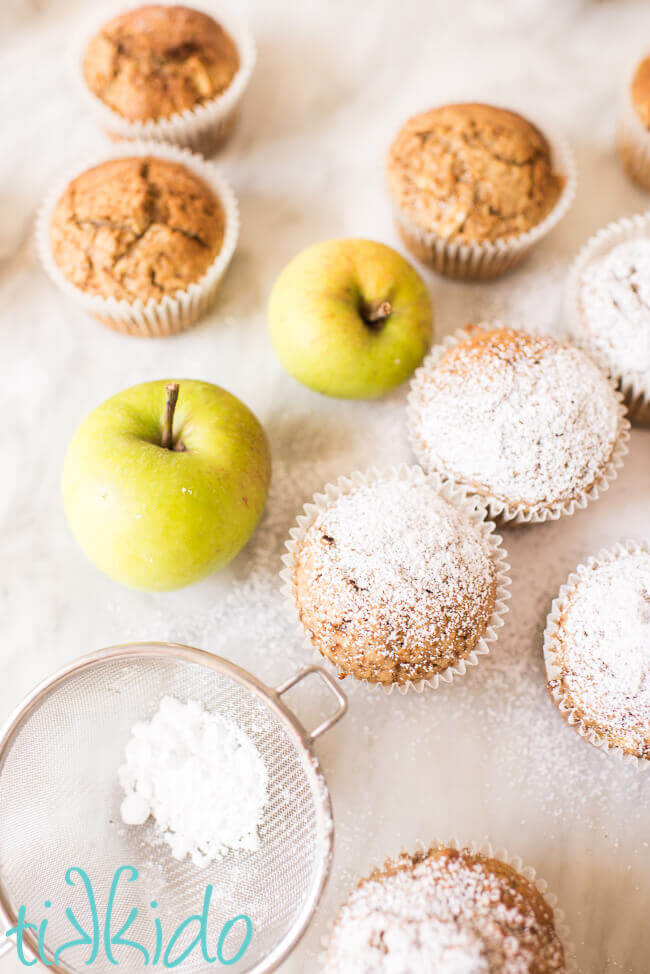 apple muffins being dusted with powdered sugar on a white marble surface.