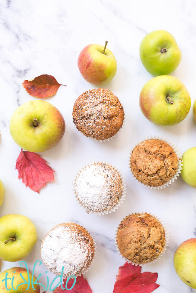 Apple muffins surrounded by fresh apples and fall leaves on a white marble surface.
