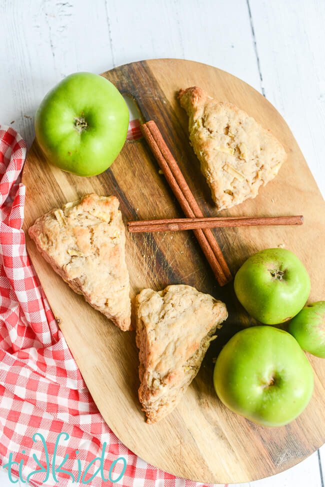 Three apple scones on a wooden cutting board, surrounded by cinnamon sticks, fresh green apples, and a red and white gingham kitchen towel.