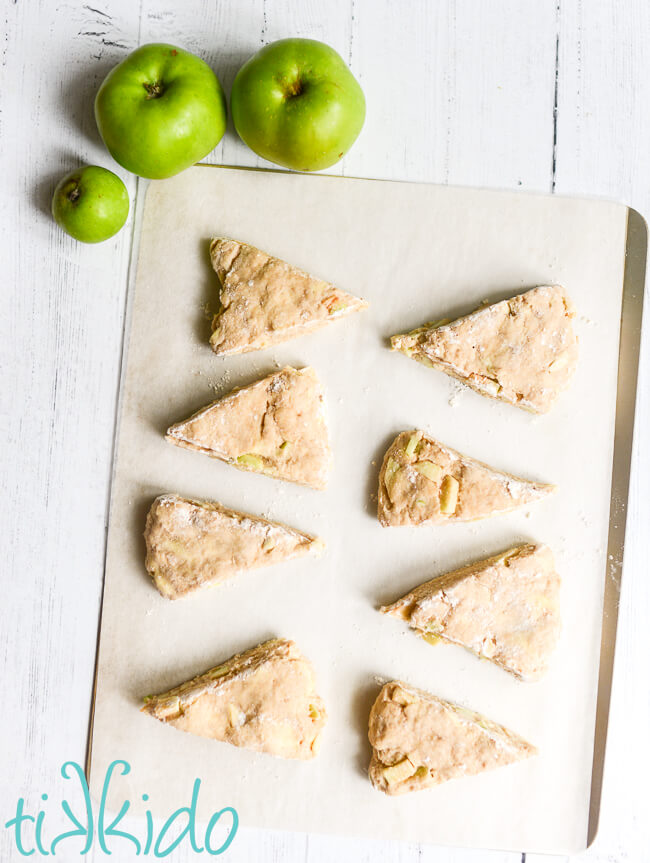 Unbaked apple scones on a parchment paper lined baking sheet, sitting on a white wooden surface next to three green apples.