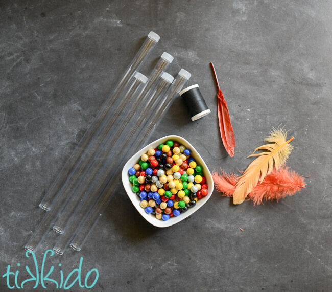 Five long and narrow clear plastic tubes, a spool of black thread, three feathers, and a small white bowl full of round, colorful chocolates.