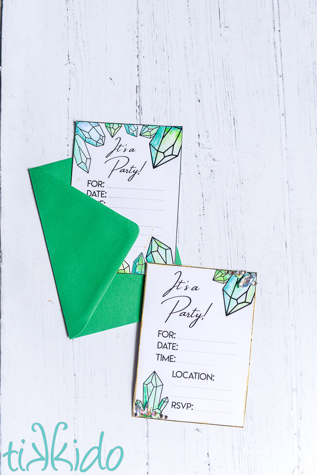 Photo of two printable, crystal-themed invitations, painted with green watercolor paints, and embellished with Czech Preciosa rhinestones.   The top invitation is nestled partially in an emerald green envelope.  Both sit on a white wooden surface.