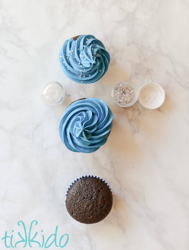 three cupcakes in stages of being iced with blue frosting and sprinkled with edible silver stars