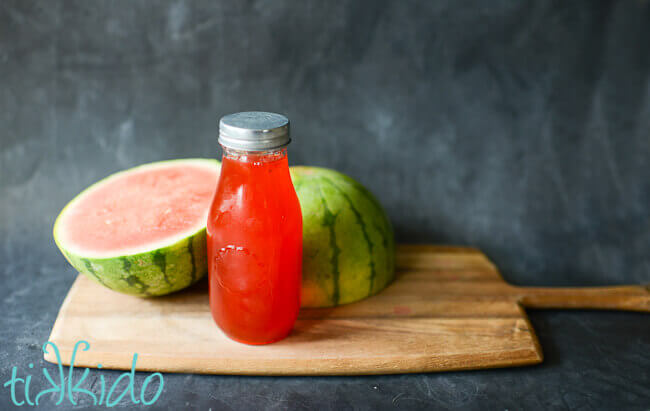 Homemade watermelon syrup in a bottle in front of a watermelon cut in half.