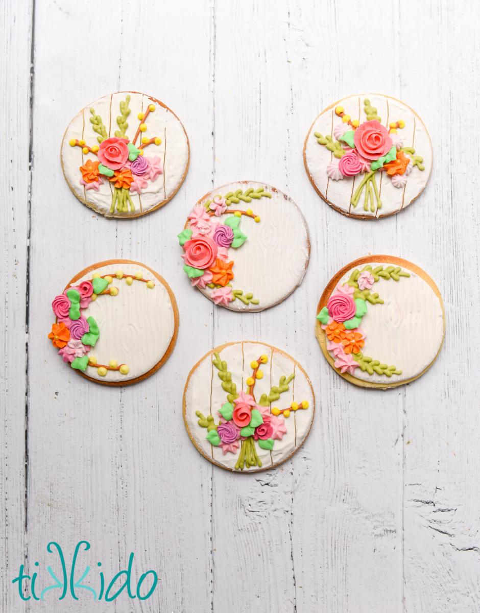 Six round sugar cookies with a rustic white wooden effect and a bouquet of spring royal icing flowers.