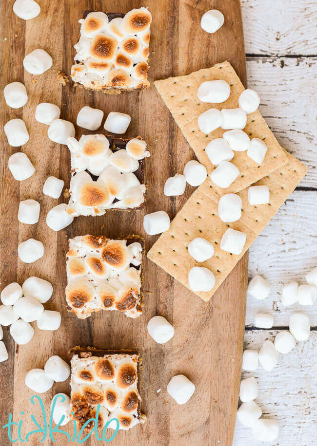 No bake s'mores bar squares on a wooden cutting board surrounded by marshmallows and graham crackers.