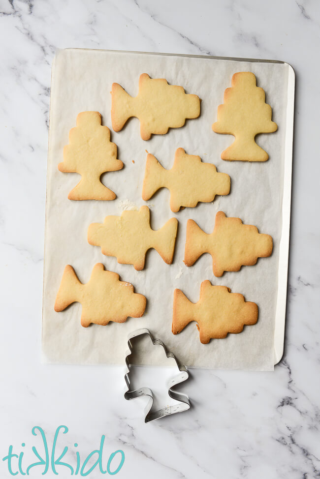 Cookie sheet lined with parchment paper, with freshly baked sugar cookies shaped like tiered cakes.  The cookie same cookie cutter rests at the bottom of the picture.