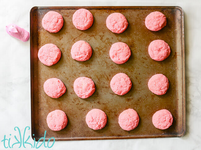 Baked pink sugar cookies on a cookie sheet on a white marble background, next to a pin Peeps candy.