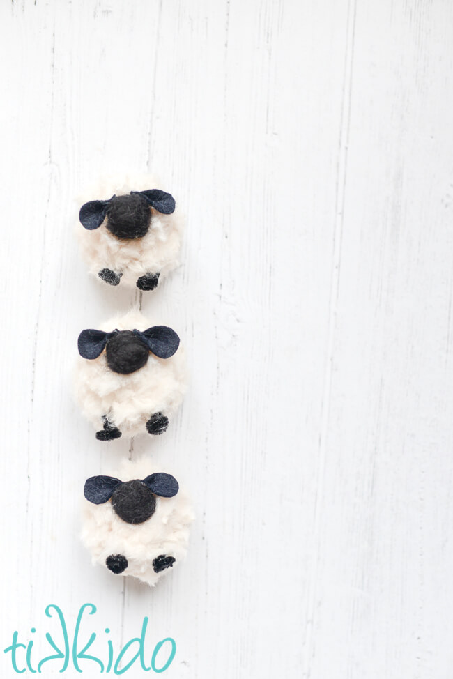 Three Pom Pom Sheep lined up vertically in a row on a white wooden surface.