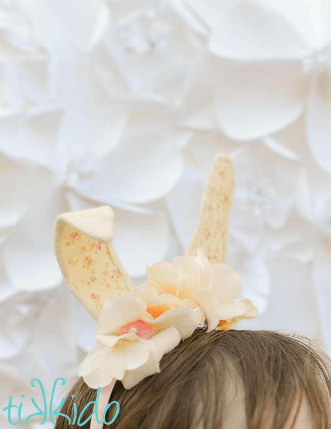 Girl wearing Bunny ear headband made with cream colored felt, a peaches and cream colored calico fabric, and ivory silk flowers on a pink headband, in front of a white surface.
