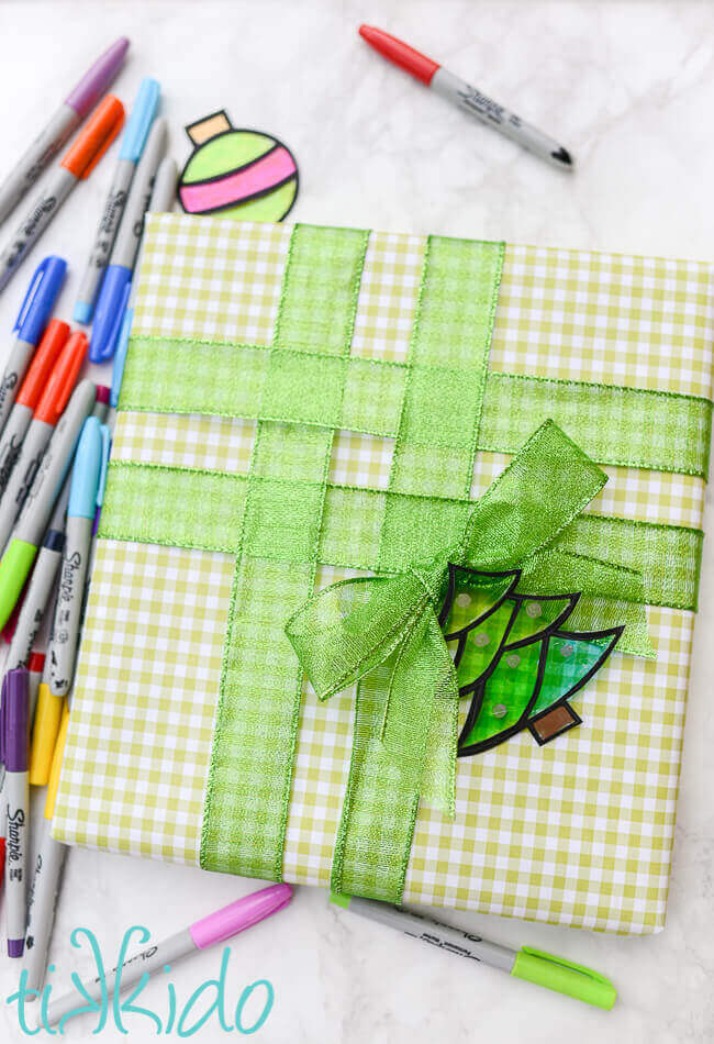 Tree shaped green faux stained glass gift tag on a present wrapped in green gingham and green ribbon. Sharpie markers surrounding present on white marble surface.
