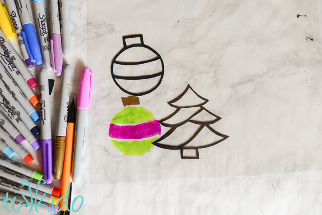 Paper outlines of a tree and ornament shape, green and pink ornament shape colored in on clear plastic film.  Sharpie markers to the left.