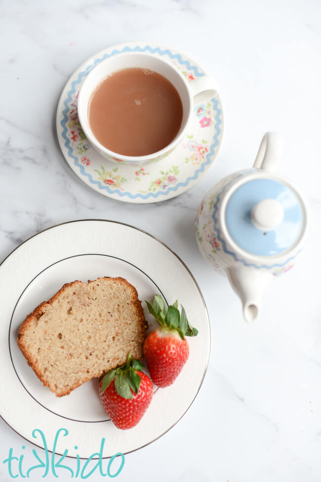 Slice of strawberry bread and two fresh strawberries on a white plate, next to a small teapot and cup of tea.