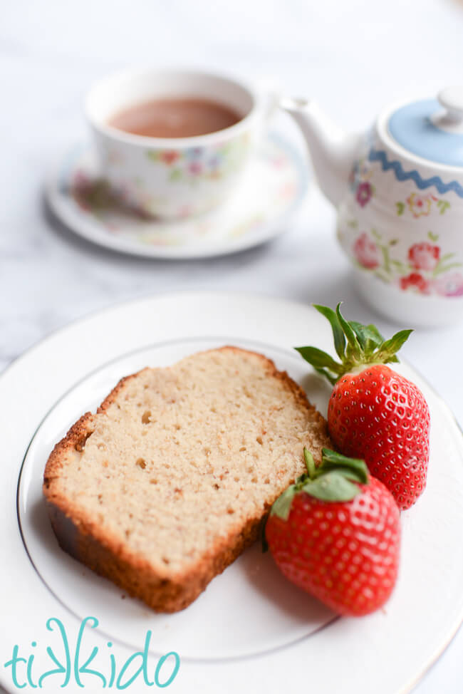 Slice of strawberry bread on a plate with two fresh strawberries, next to a tea pot and cup of tea.