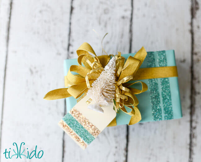 Christmas present wrapped in aqua and gold, topped with a bottle brush tree and a DIY Gift Tag