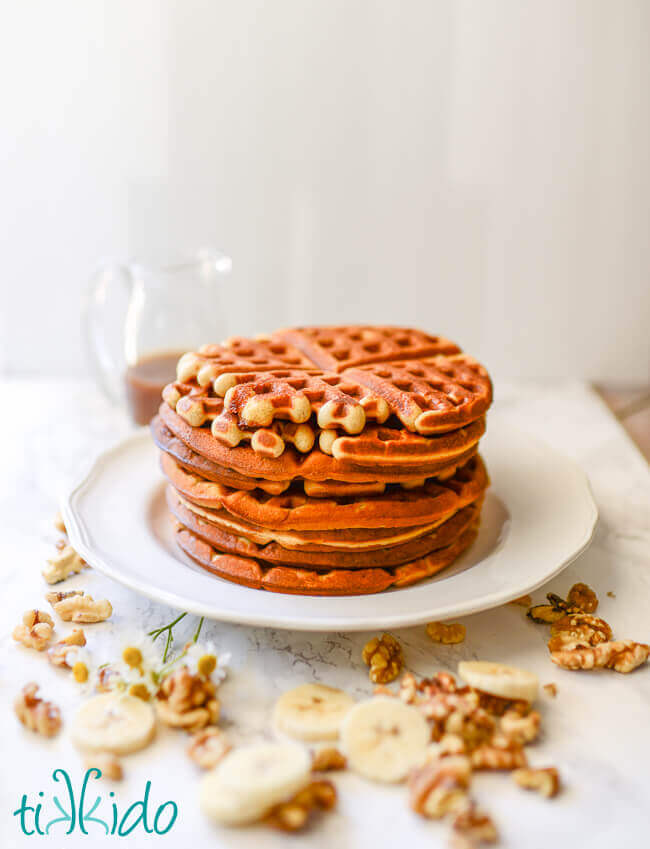 Stack of banana bread waffles on a white plate on a white background, with slices of bananas and walnuts around the plate.