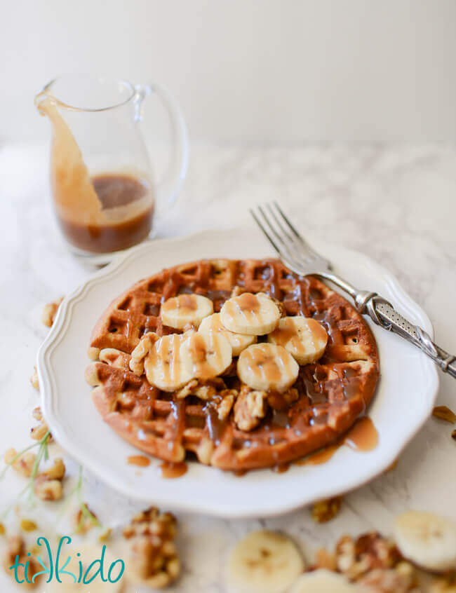 Banana bread waffles topped with sliced bananas, walnuts, and brown sugar caramel syrup on a white plate with a fork, small pitcher of syrup above the plate, and scattered banana slices and walnuts around the plate.