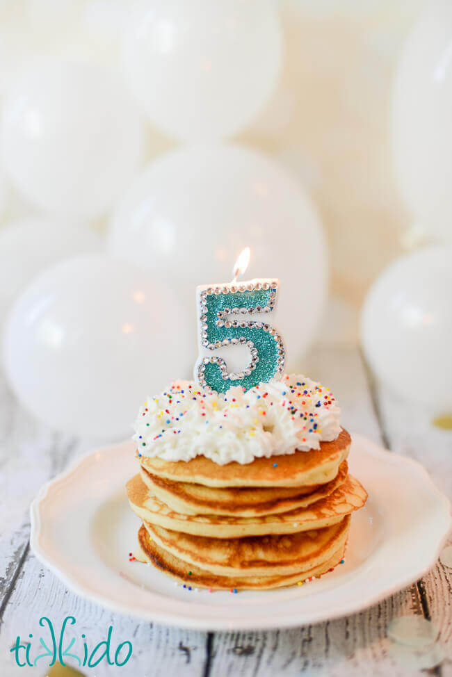 Stack of birthday cake pancakes with whipped cream, sprinkles, and a number 5 candle on top.