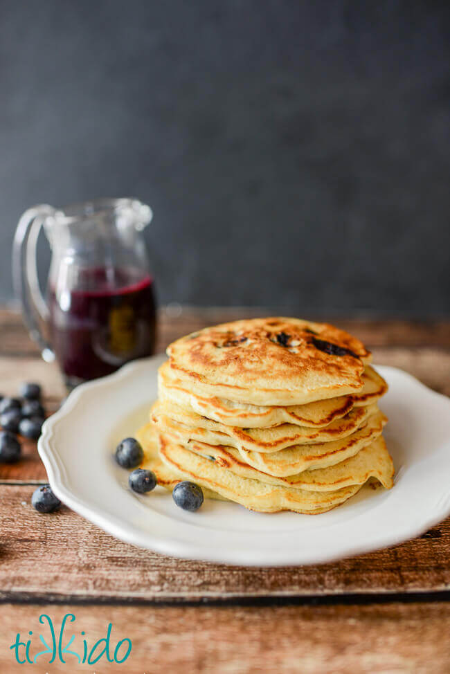 Stack of blueberry buttermilk pancakes with a pitcher of blueberry syrup.