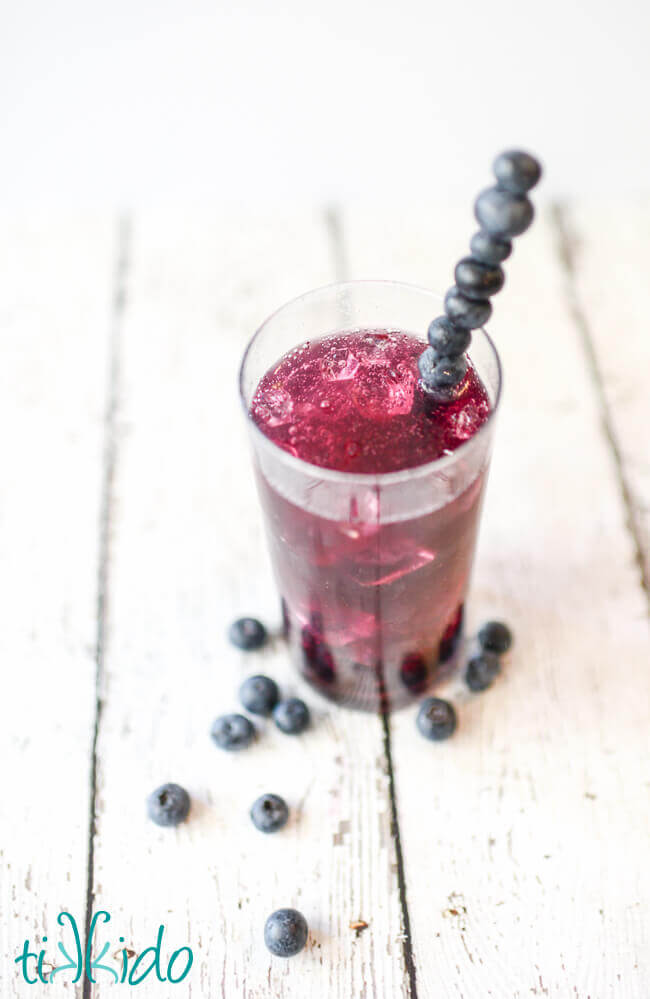 Tall glass of a vibrant bluish-purple blueberry soda with ice, with a stirrer made from skewered blueberries, on a white surface with fresh blueberries scattered around.