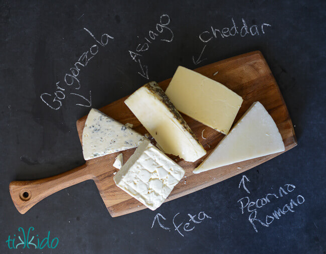 Wooden cutting board holding five kinds of cheese, on black chalkboard background.