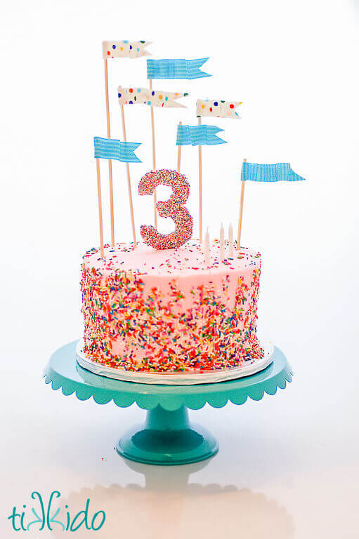 Sprinkled covered cake with a sprinkle covered 3 cake topper, and blue striped and multi colored polka dot fabric flag cake toppers that are posable and look like they're blowing in the wind.