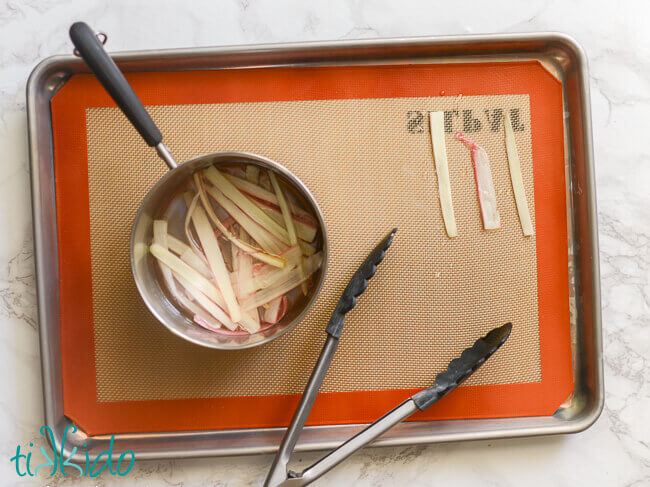 Small saucepan with strips of rhubarb and sugar water, on a sheet pan lined with a silpat, tongs arranging the strips of rhubarb on the silpat.