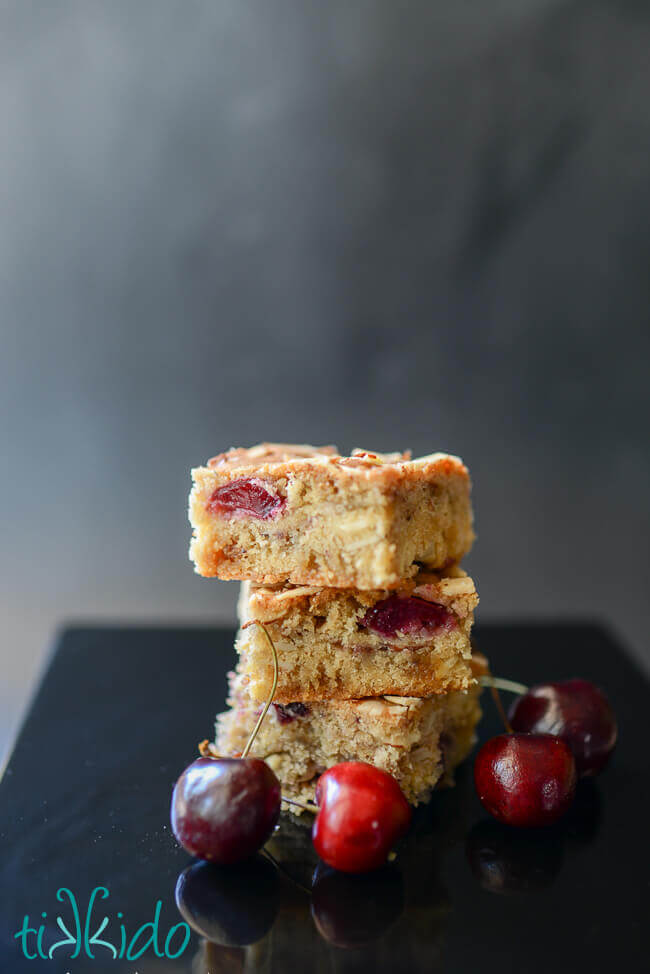 Three cherry almond bar cookies stacked, on a black surface, surrounded by fresh cherries.
