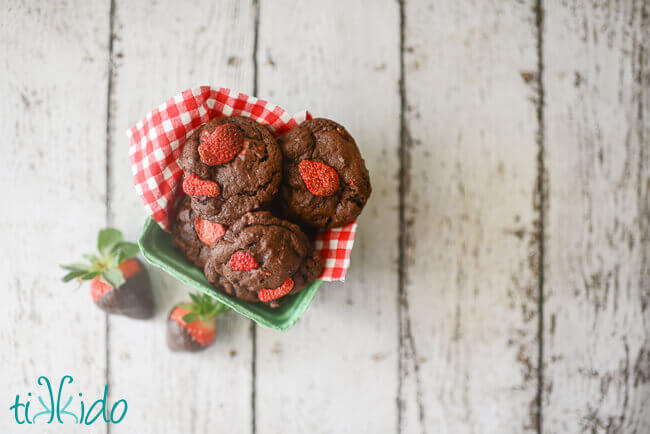 Chocolate Covered Strawberry Cookies in a strawberry punnet on a white, weathered wooden surface.  Two fresh chocolate covered strawberries sit next to the basket of cookies.