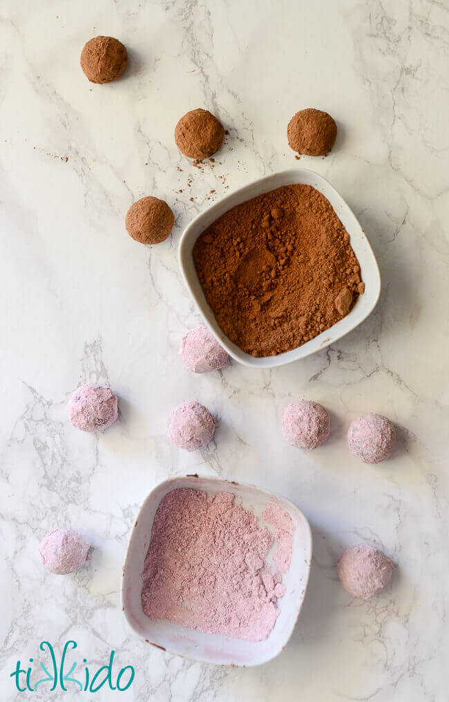 Truffles rolled in cocoa powder and in raspberry powdered sugar.  Bowls of coatings beside the truffles.  White marble background.