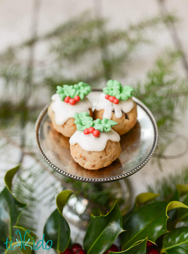 Three Figgy Pudding Cookies on a small silver stand, surrounded by fresh holly and evergreen branches.