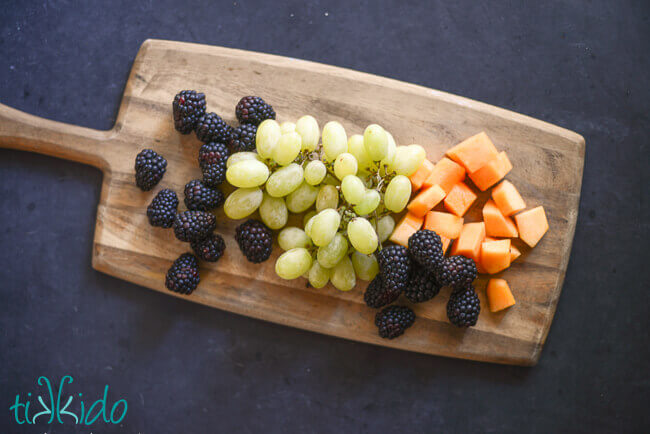 Blackberries, green grapes, and cantaloupe melon on a wooden cutting board.