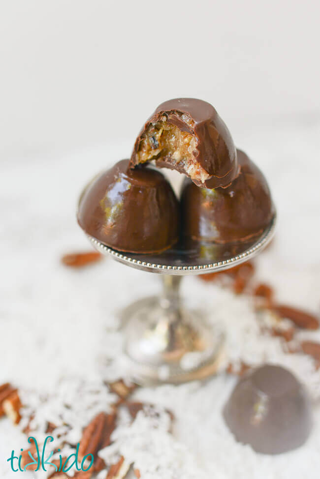 Chocolate bon bons on a small metal serving stand, with coconut and pecans scattered around base. One bite out of the top chocolate, exposing the german chocolate filling.