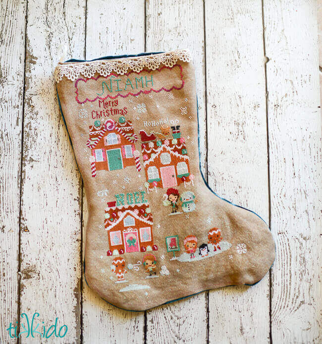 Cross stitch gingerbread house Christmas stocking on a weathered white wooden background.