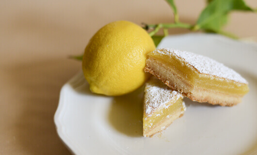 Two classic lemon bars cut into a diamond shape on a white plate, next to a fresh lemon with leaves attached to the stem.